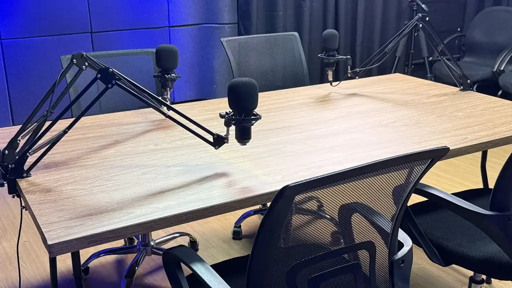 Detalhe da mesa de podcast da OFBOX com microfones montados em braços articulados e cadeiras ao redor.