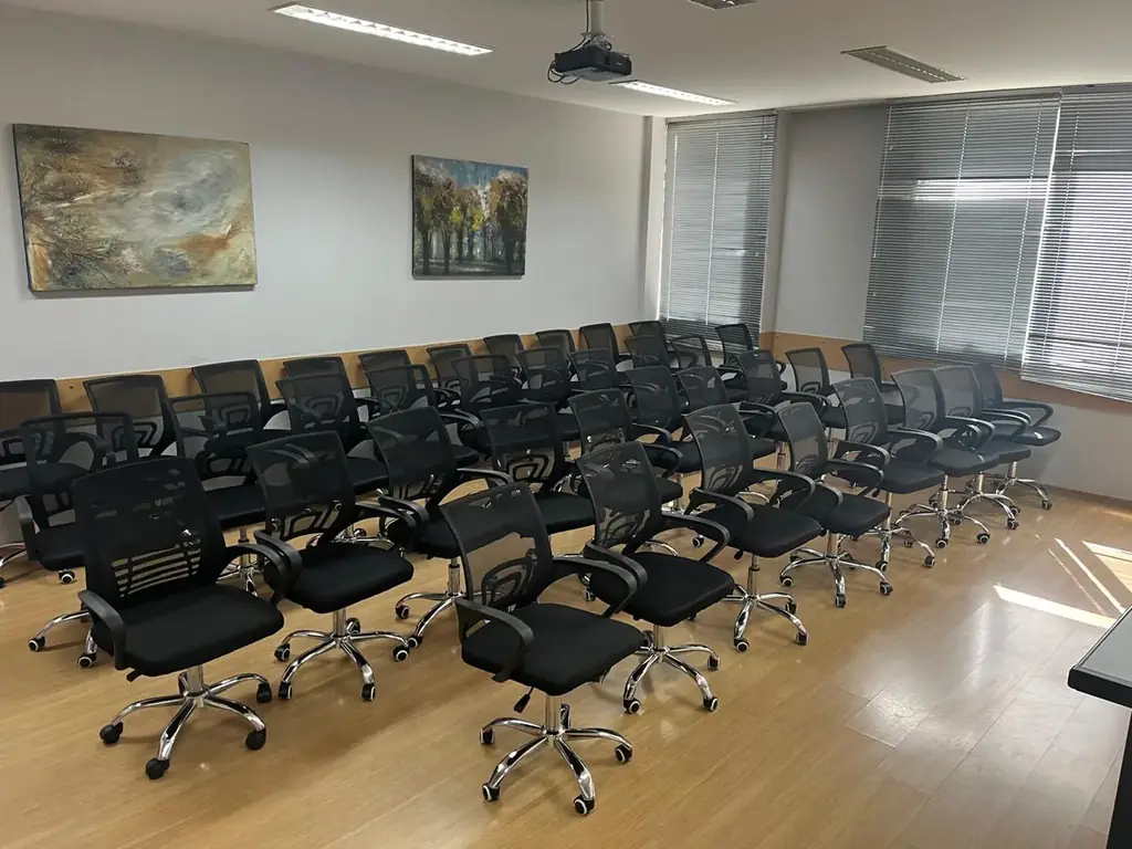 Vista ampla da sala de aula OFBOX com cadeiras giratórias organizadas em fileiras, projetor no teto e iluminação profissional.