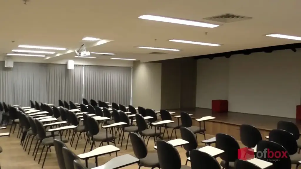 Auditório OFBOX com cadeiras universitárias enfileiradas voltadas para palco em madeira, cortinas claras, projetor de teto e púlpito vermelho.