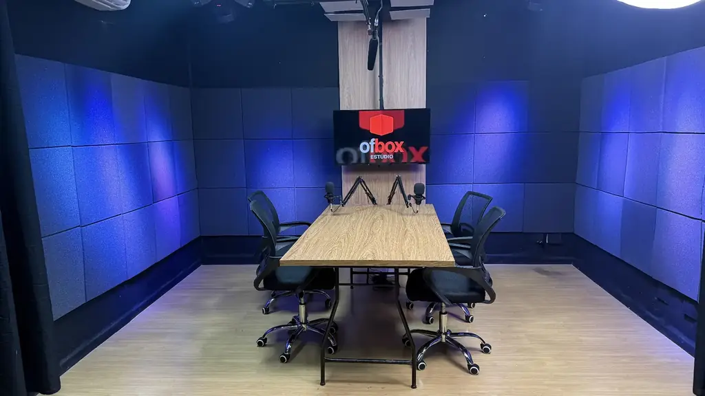 Vista frontal do estúdio OFBOX com iluminação azul, mesa de gravação e quatro posições para participantes.