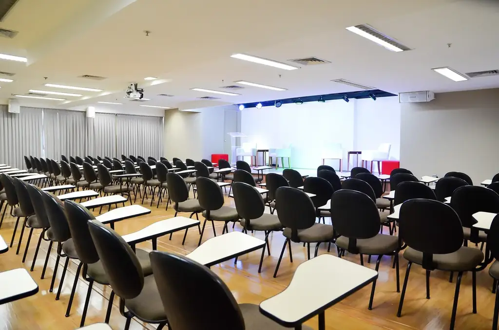 Auditório OFBOX com fileiras de cadeiras com prancheta voltadas para palco iluminado, púlpito de acrílico e iluminação cênica azul.