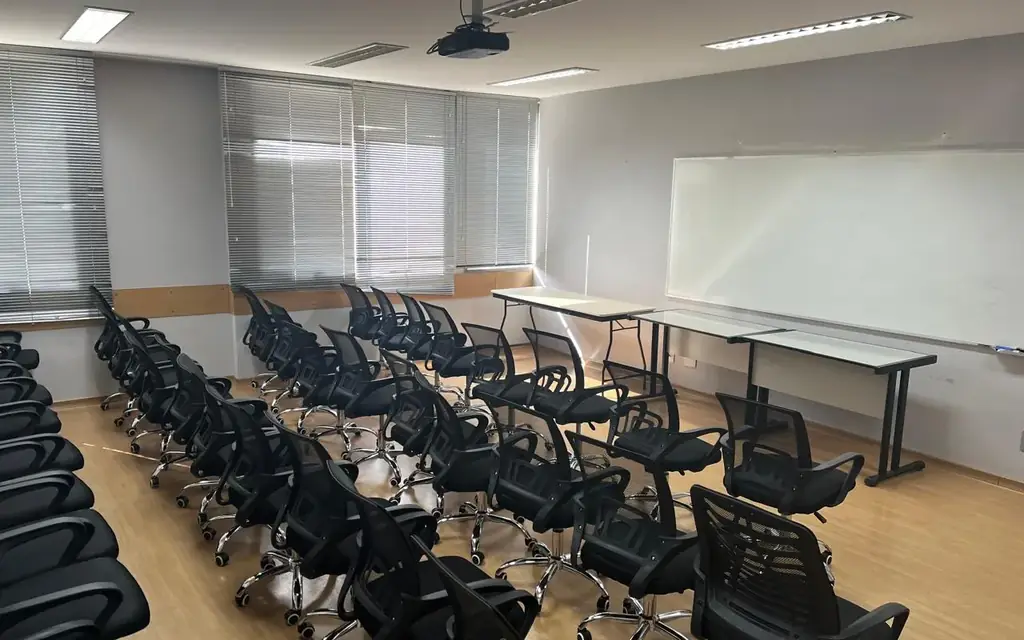 Sala de aula OFBOX com cadeiras giratórias pretas em fileiras, quadro branco na parede, mesas de apoio e projetor suspenso no teto.