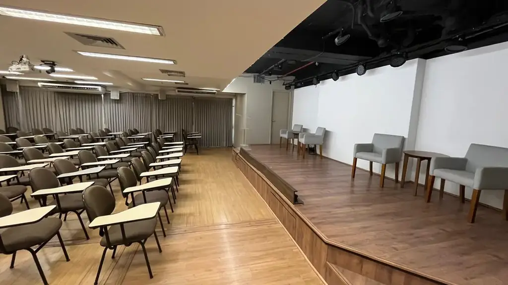 Auditório OFBOX visto da plateia com palco elevado em madeira, parede branca para projeção, poltronas e iluminação cênica de teto preto.